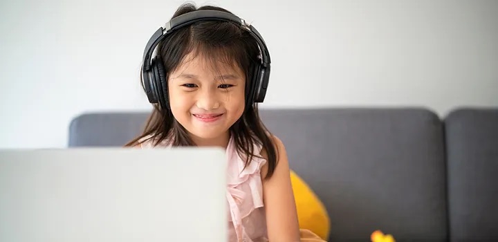 young girl smiling and looking at laptop and wearing headphones and sitting on grey couch
